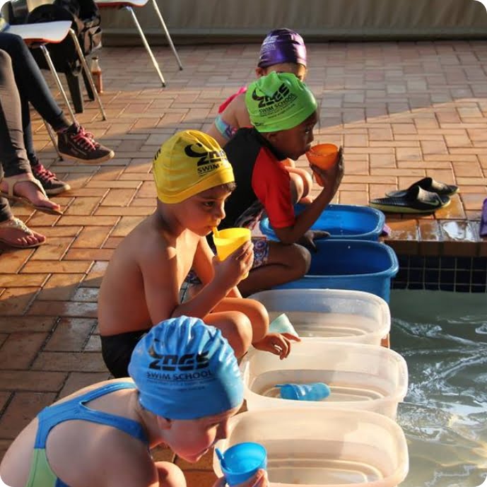 Children in Zogs Swim School caps sitting poolside, sipping drinks and soaking their feet after a swim session.