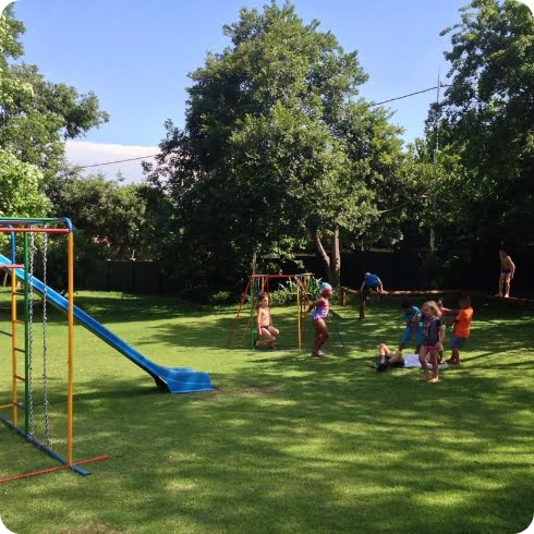Children playing on swings and a slide in the secure outdoor play area at Zogs Swim School, surrounded by trees and greenery.